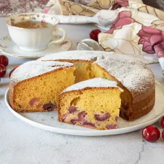 Una fetta di torta alle ciliegie, soffice e profumata, cosparsa di zucchero a velo. Ciliegie fresche e una tazza di tè completano la scena.