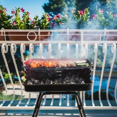 Grill z mięsem i warzywami na balkonie, w tle kwiaty i błękitne niebo.