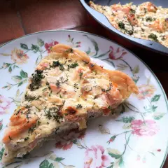 Pyszna tarta z łososiem i porem, posypana świeżym koperkiem, czeka na talerzu w kwiaty. Obok w naczyniu reszta wypieku.