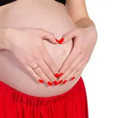 Mani con unghie rosse formano un cuore sul pancione. Si possono fare le unghie in gravidanza, con cura e attenzione.