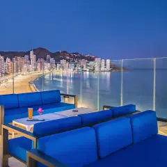 Sofás azules en terraza con vistas a la playa y la ciudad iluminada de Barceló Benidorm Beach al anochecer.