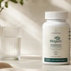 Flacone di Magtein, integratore alimentare di magnesio treonato, accanto a un bicchiere d'acqua e una pianta.