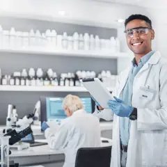 Uśmiechnięty technik farmacji w okularach ochronnych trzyma tablet w laboratorium, gdzie na półkach stoją liczne butelki, a w tle pracuje inna osoba.