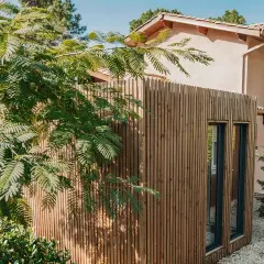 Extension de maison en bois moderne, bardage vertical, fenêtres sombres. Un espace de vie supplémentaire sans permis de construire, intégré dans un jardin verdoyant.