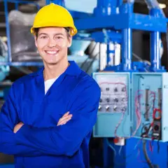 Uśmiechnięty pracownik w żółtym kasku i niebieskim kombinezonie, stojący przed panelem elektrycznym. To może być pracownik hurtowni elektrotechnicznej Comel Rumia.