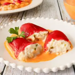 Pimientos rellenos de bacalao, cubiertos en una cremosa salsa naranja, servidos en un plato blanco con un tenedor dorado al lado.