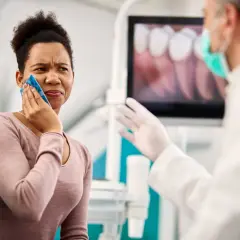 Mujer con compresa fría en la mejilla, sufriendo un alveolitis con dolor insoportable, mientras el dentista le explica algo.