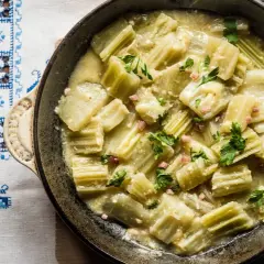 Cardo en salsa cremosa con trocitos de jamón y perejil, servido en sartén sobre mantel bordado. Un plato delicioso con cardo y almendras.