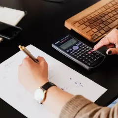 Mani che scrivono su fogli, usano una calcolatrice per lo scorporo IVA e una tastiera in legno.