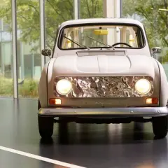 En ljusbeige Renault 16 står framför stora fönster med grönska utanför. Bilens front med sina runda strålkastare och kromade grill är i fokus.