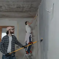 Para robi poprawki na pomalowanej ścianie. Mężczyzna z wałkiem w ręku, kobieta na drabinie maluje sufit.