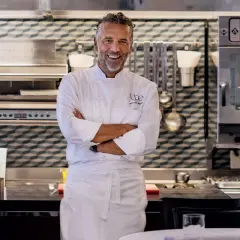 Chef Perbellini, sorridente in cucina, con le braccia conserte. Dietro di lui, attrezzature professionali e un forno acceso.