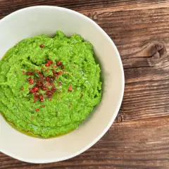 Jasnozielone puree z groszku, ozdobione różowym pieprzem i oliwą, w białej misce na drewnianym tle.