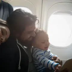 Familj flyger med bebis. Pappa håller ett leende barn som tittar på en surfplatta, mamman sover bredvid.