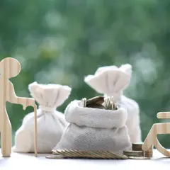 Drewniane figurki starszej osoby z laską i osoby na wózku inwalidzkim, obok worków z pieniędzmi. Jak załatwić opiekunkę do starszej osoby z mops?