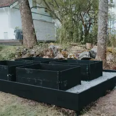 Nymålade pallkragar i svart står redo för plantering i trädgården, med ett vitt hus i bakgrunden.