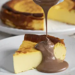 Un trozo de basque cheesecake cremoso recibe un chorro de chocolate fundido. Otro pastel entero se ve desenfocado al fondo.