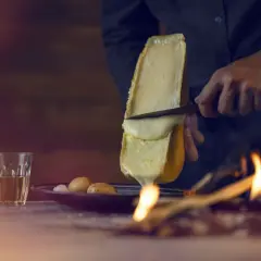 Un morceau de fromage à raclette fond sur un feu, rappelant son origine alpine. Des pommes de terre et un verre de vin complètent la scène.