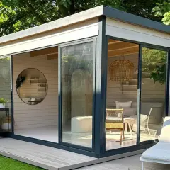 Modernes Gartenhaus mit Glasfront, das zum Streichen einlädt. Gemütliche Inneneinrichtung mit Rattan-Akzenten und Liegestuhl im Freien.