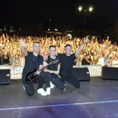 Trzech muzyków na scenie, z gitarą i mikrofonem, pozuje przed rozentuzjazmowanym tłumem. To z pewnością udane imieniny Jana!