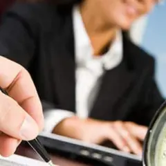 A hand holds a pen, poised to take notes, as a person in a suit works at a laptop. This scene evokes the process of creating board meeting minutes examples.