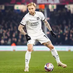 Piłkarz w białej koszulce z tatuażami na rękach i nogach, gdzie gra puchacz, przygotowuje się do strzału na zielonej murawie stadionu.