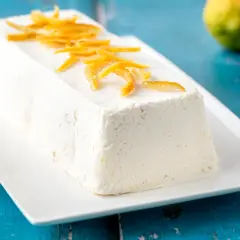 Delizioso semifreddo al limone, guarnito con scorzette candite, su un piatto bianco. Due limoni freschi sullo sfondo.