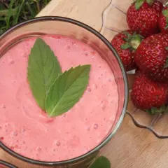 Pyszny, różowy shake truskawkowy z listkami mięty, obok świeże truskawki na talerzyku. Idealny na letnie orzeźwienie.