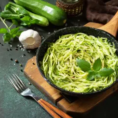 Pyszny makaron keto z cukinii w patelni, udekorowany bazylią. Obok świeże cukinie, czosnek i przyprawy.