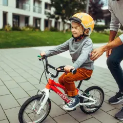 Chłopiec w kasku uczy się jeździć na czerwonym rowerze, a tata go asekuruje. To świetny przykład, jak dobrac rower dla dziecka.