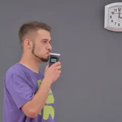 Młody mężczyzna dmucha w alkomat, sprawdzając, ile promili ma po lampce szampana. Zegar na ścianie pokazuje czas.