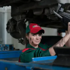 Mécanicienne sous une voiture, le visage barbouillé de cambouis. Elle est experte automobile, mais son regard suggère un abus de pouvoir.