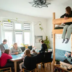 Rodzina gra w planszówkę w domu pomocy społecznej "Przystań". Dzieci siedzą na łóżku piętrowym, obserwując grę.