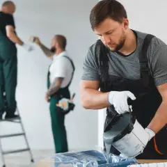 Malowanie ścian w bloku. Dwóch mężczyzn w kombinezonach przygotowuje się do pracy, jeden nalewa farbę, drugi jest na drabinie.