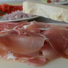 Cieniutkie plastry szynki crudo, obok kawałek parmezanu i talerz z posiekanymi pomidorami i rozmarynem.