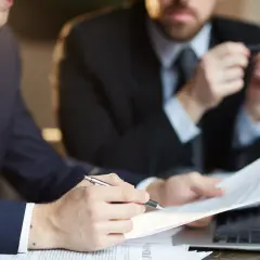 Dwóch mężczyzn w garniturach analizuje dokumenty przy laptopie.