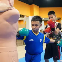 A young boy in a blue shirt practices a boxing feint, throwing a punch at a training dummy while other children watch.