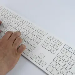 Dłonie piszą na białej klawiaturze laptopa. Widoczny jest opis klawiszy, w tym litery i symbole.