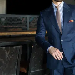 Elegancki mężczyzna w granatowym garniturze, z niebiesko-białą koszulą i brązowym krawatem. W kieszeni marynarki widoczny kolorowy poszetka. Dyskretny dekolt koszuli podkreśla stylizację.