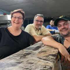Fröhliche Menschen am holzmarkt ilsfeld. Eine Frau mit Brille und schwarzem Shirt, ein Mann mit gelbem Hemd und ein Mann mit Kappe lächeln in die Kamera.