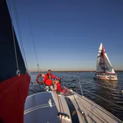 Dwóch żeglarzy na pokładzie, wykonujących zwrot przez rufę. Na wodzie widać inne łodzie.