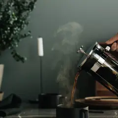 Här hälls nybryggt kaffe till kaffepressen i en mörk mugg. Ångan stiger från den varma drycken.