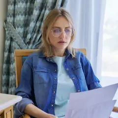 Kobieta w okularach z uwagą czyta dokumenty. Zastanawia się, czy można wysłać list bez nadawcy, gdy na stole stoi szklanka wody.