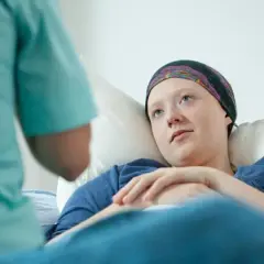 Młoda kobieta w chustce na głowie leży w łóżku szpitalnym, rozmawiając z pielęgniarką. Długie zwolnienie po operacji oszczędzającej jest nieuniknione.
