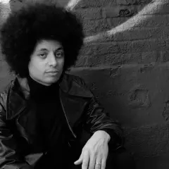 Jose James z burzą afro, w skórzanej kurtce i golfie, siedzi na tle ściany z graffiti.
