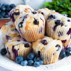 Stos świeżo upieczonych muffinek z borówkami, polanych lukrem, z kilkoma borówkami na talerzu.