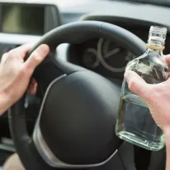 Ręce kierowcy na kierownicy, jedna trzyma butelkę alkoholu. To niebezpieczne zachowanie, które łamie limit alkoholu w Polsce.