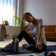 Erschöpfte Frau sitzt auf einer Matte, Hand an der Stirn. Vielleicht ein Fehler beim Muskelaufbau? Trainingsgeräte liegen herum.