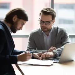 Dwóch mężczyzn w garniturach analizuje dokumenty. Jeden wskazuje palcem na umowę o dzieło po angielsku, drugi podpisuje.