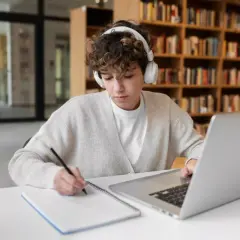 Młoda osoba w słuchawkach uczy się w bibliotece, notując na kartce. Czy nauczanie domowe jest płatne?
